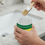 A person holds a clear jar of urethane waterproof coating with a green and yellow label while dipping a paintbrush into the container. The background shows a blurred kitchen sink and countertop.