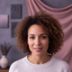 A happy, diverse woman in her late 20s to early 30s smiles gently, looking at the viewer. Blurred dusty rose and soft lavende